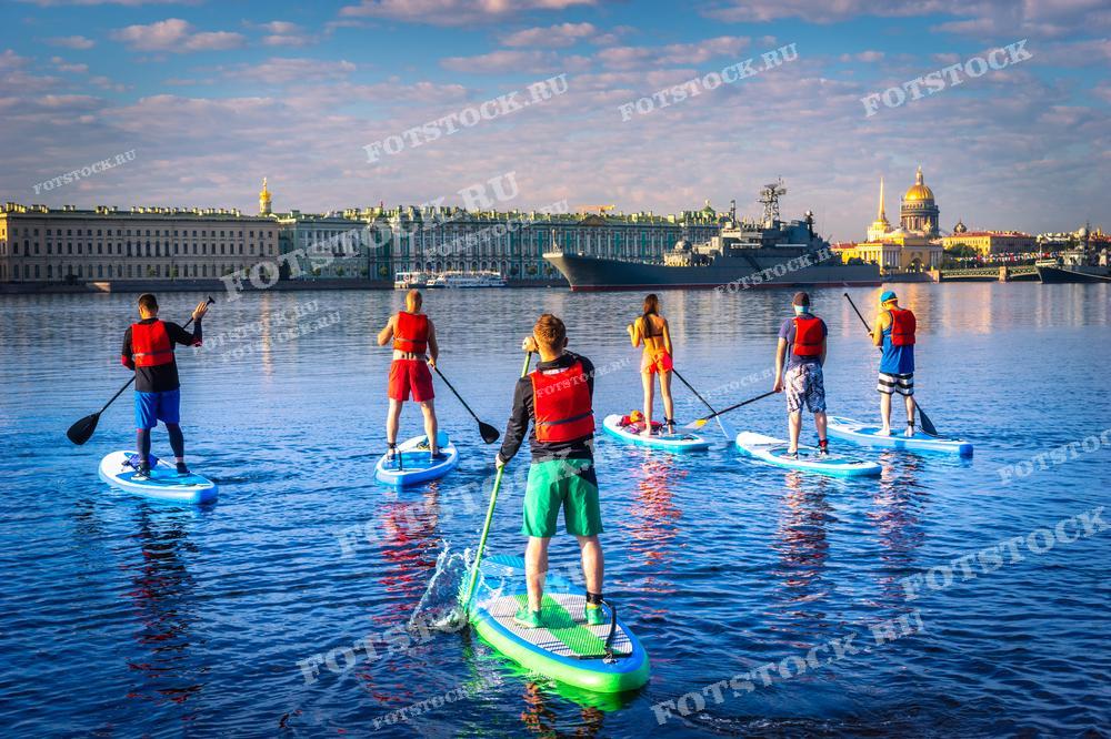На этой фотографии группа людей занимается стендап паддлингом (SUP) на фоне исторического центра Санкт-Петербурга. Гребцы на SUP-досках равномерно распределены по водной глади Невы, создавая красочную и динамичную сцену. Вдалеке видны Зимний дворец и купола Исаакиевского собора под ясным утренним небом. Этот снимок отлично подойдёт для спортивных журналов, туристических каталогов, промо-материалов об активном отдыхе на воде или публикаций о здоровом образе жизни.